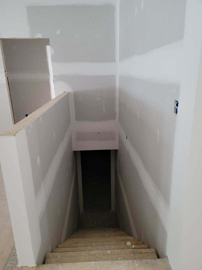 Wood staircase with white risers ascending to a dimly lit room, smooth plaster walls, light-colored flooring, and a closed door visible at the top.
