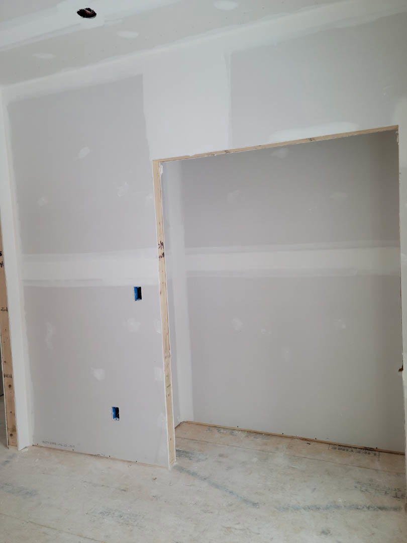 White plaster wall with a rectangular hole, wooden door frame, light-colored flooring, and partial view of a bathtub in a remodeled interior room