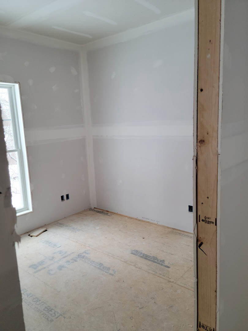 Unfinished room with white plaster walls, large window framed in wood, concrete floor marked with construction notes, and exposed wood plank featuring a central hole.