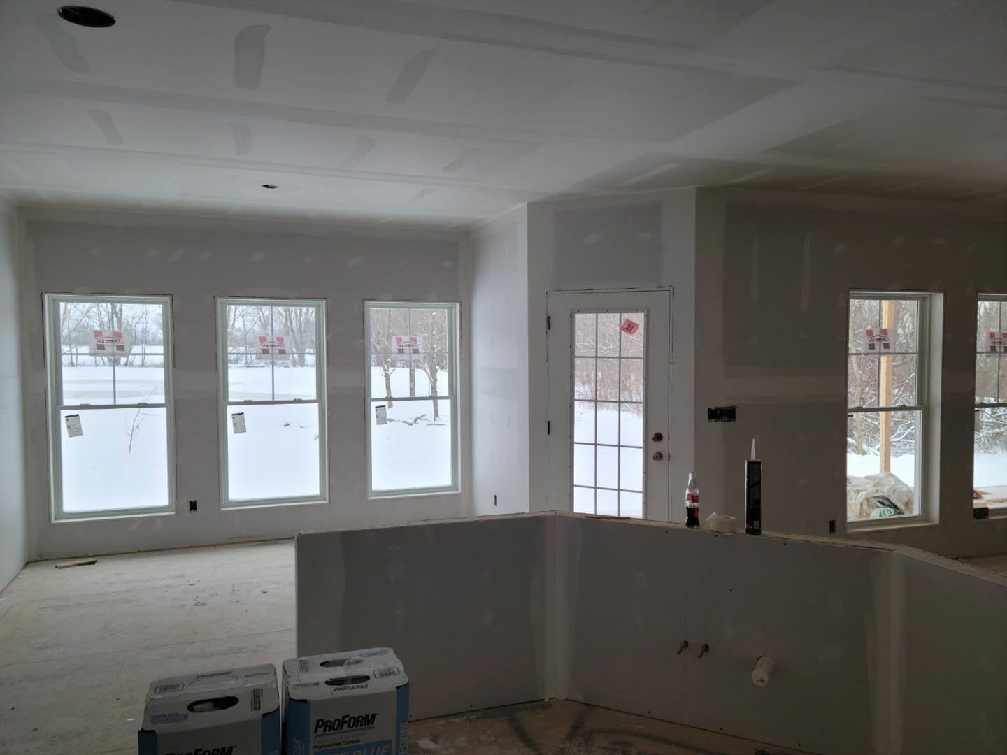 Bright room with white plaster walls, large windows, and a door featuring a window; boxes with labels are placed near the wall, and a sign is visible on one window.