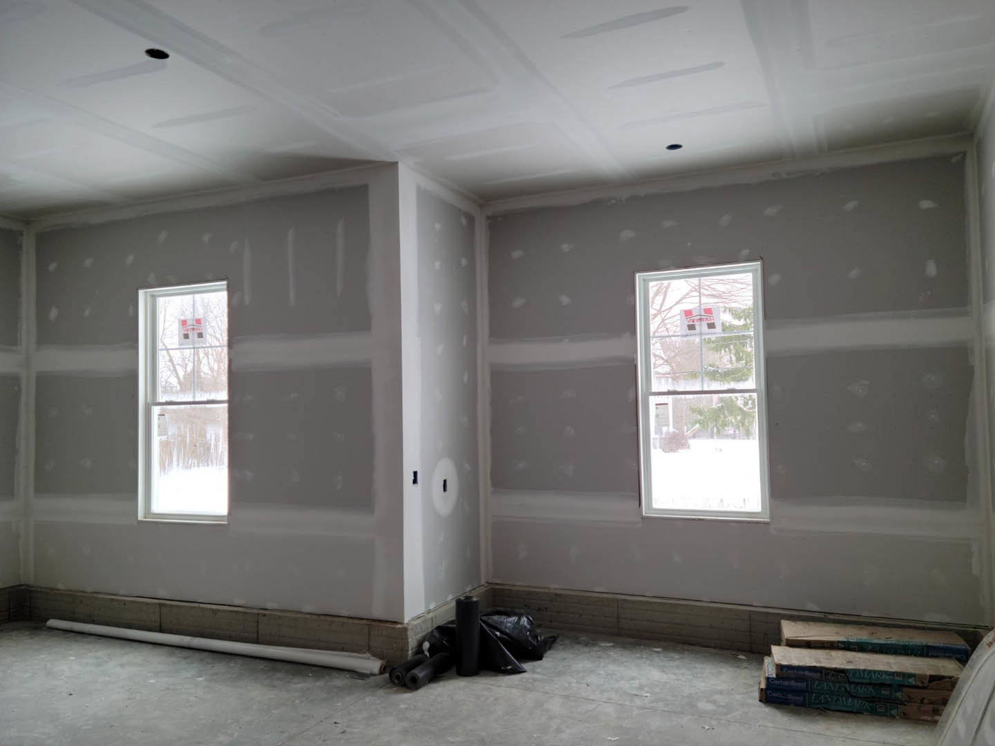 Bright room with two large windows, plaster walls, light wood flooring, stack of books on the floor, roll of black plastic near the wall, snowy landscape visible through window