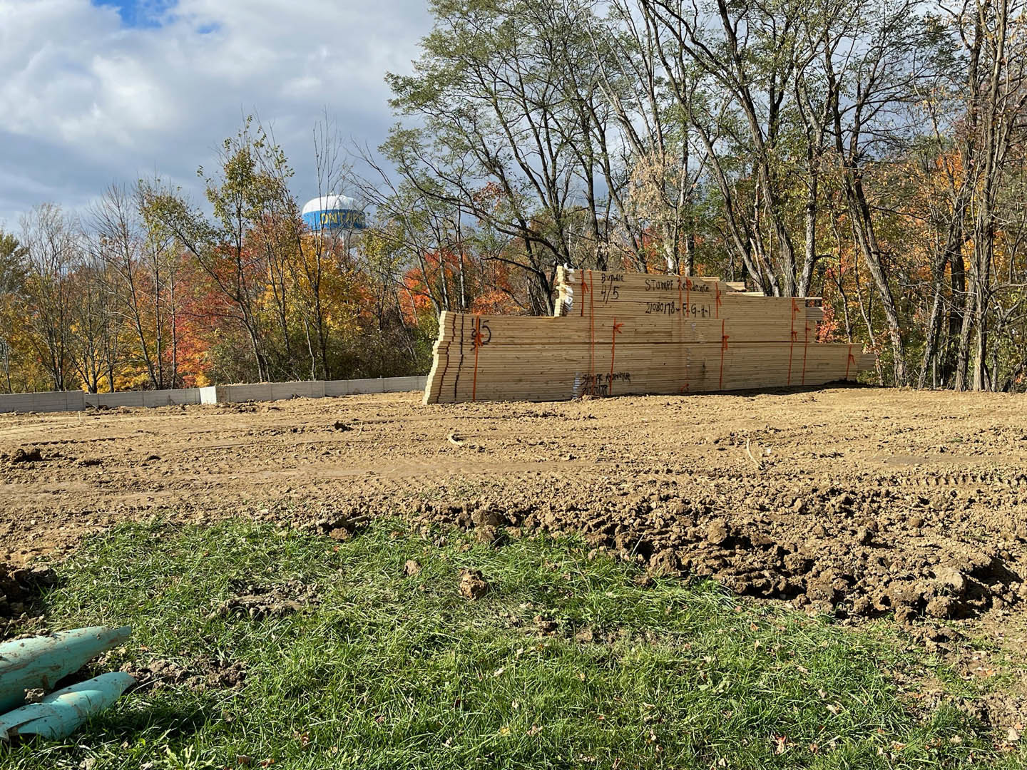 Stack of rough-cut lumber with black markings resting on bare dirt in an open grassy field, blue and white water tank visible in background under cloudy sky