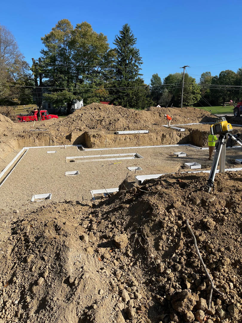 Construction site with exposed soil, foundation work in progress, construction worker near yellow and grey machinery, trees with dense foliage in background, power lines visible