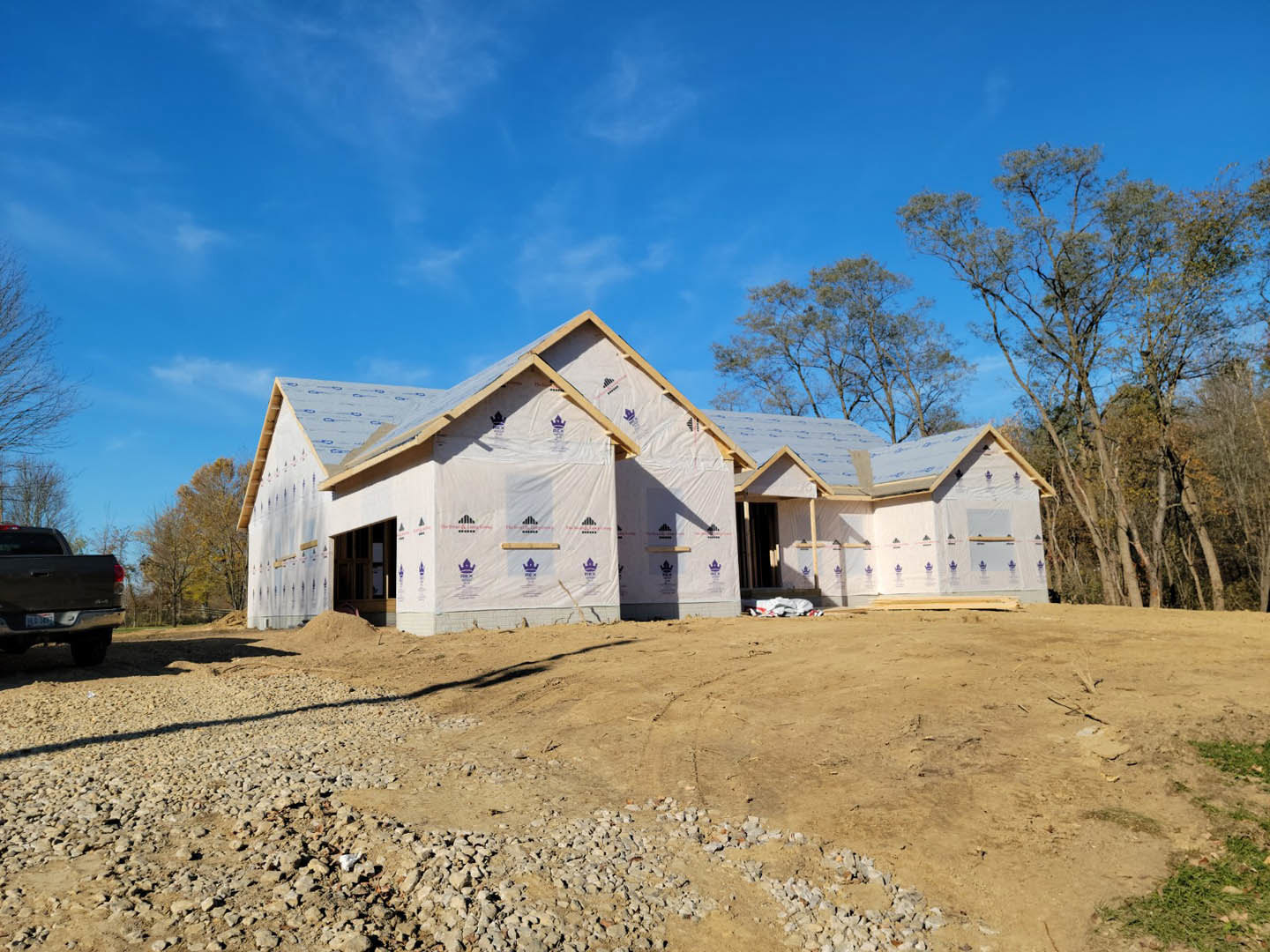 Framed house under construction with exposed wood, plastic sheeting, gravel pile, and surrounding trees beneath a clear blue sky