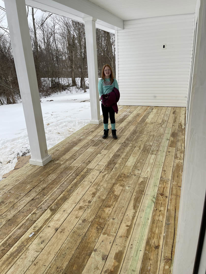 Woman wearing winter clothing stands on snowy wooden deck outside custom home, white siding and trees visible in background.