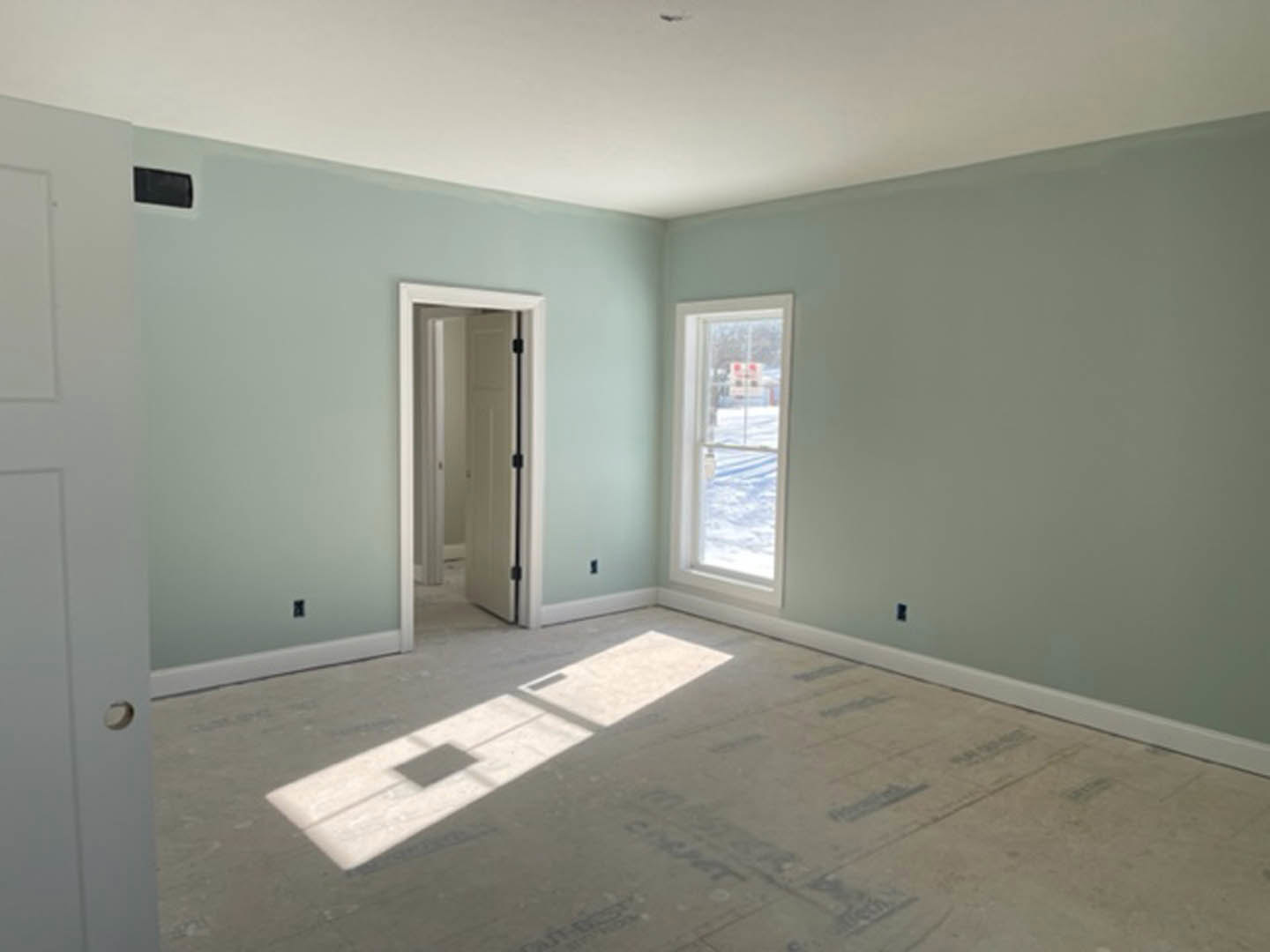 White door with black handle beside large window overlooking snowy yard, sunlight casting square shadow on light-colored floor, black rectangular object resting on white surface