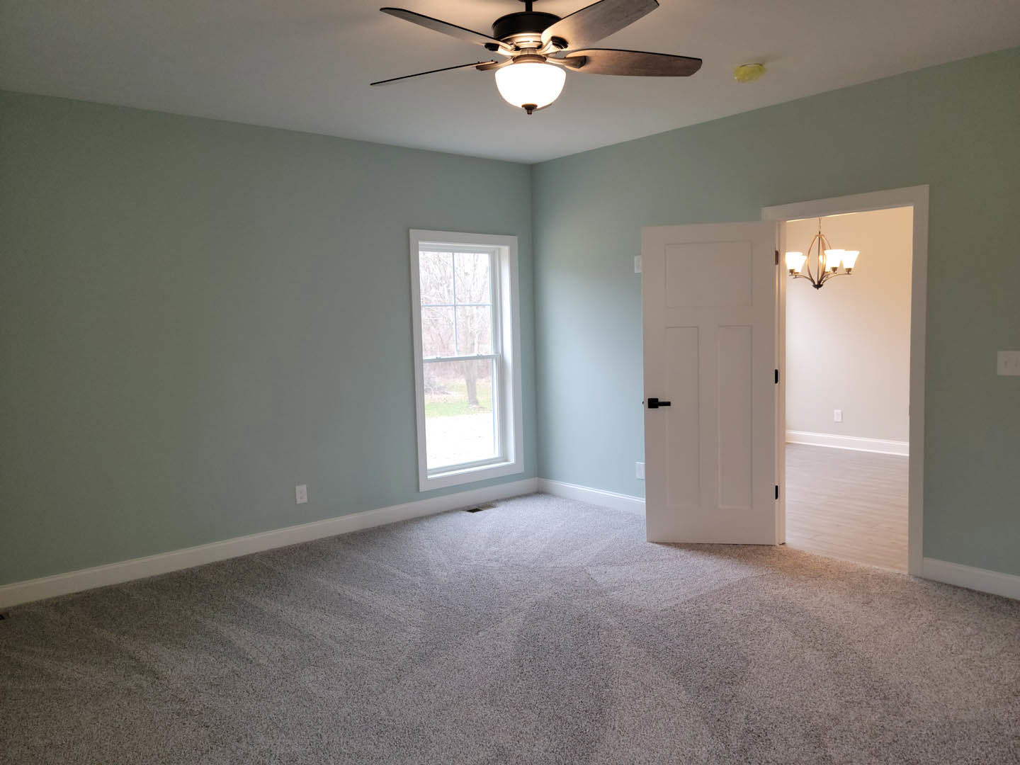 Carpeted room with white walls, ceiling fan with light, white door with black handle, window showing tree outside, wall-mounted light fixture