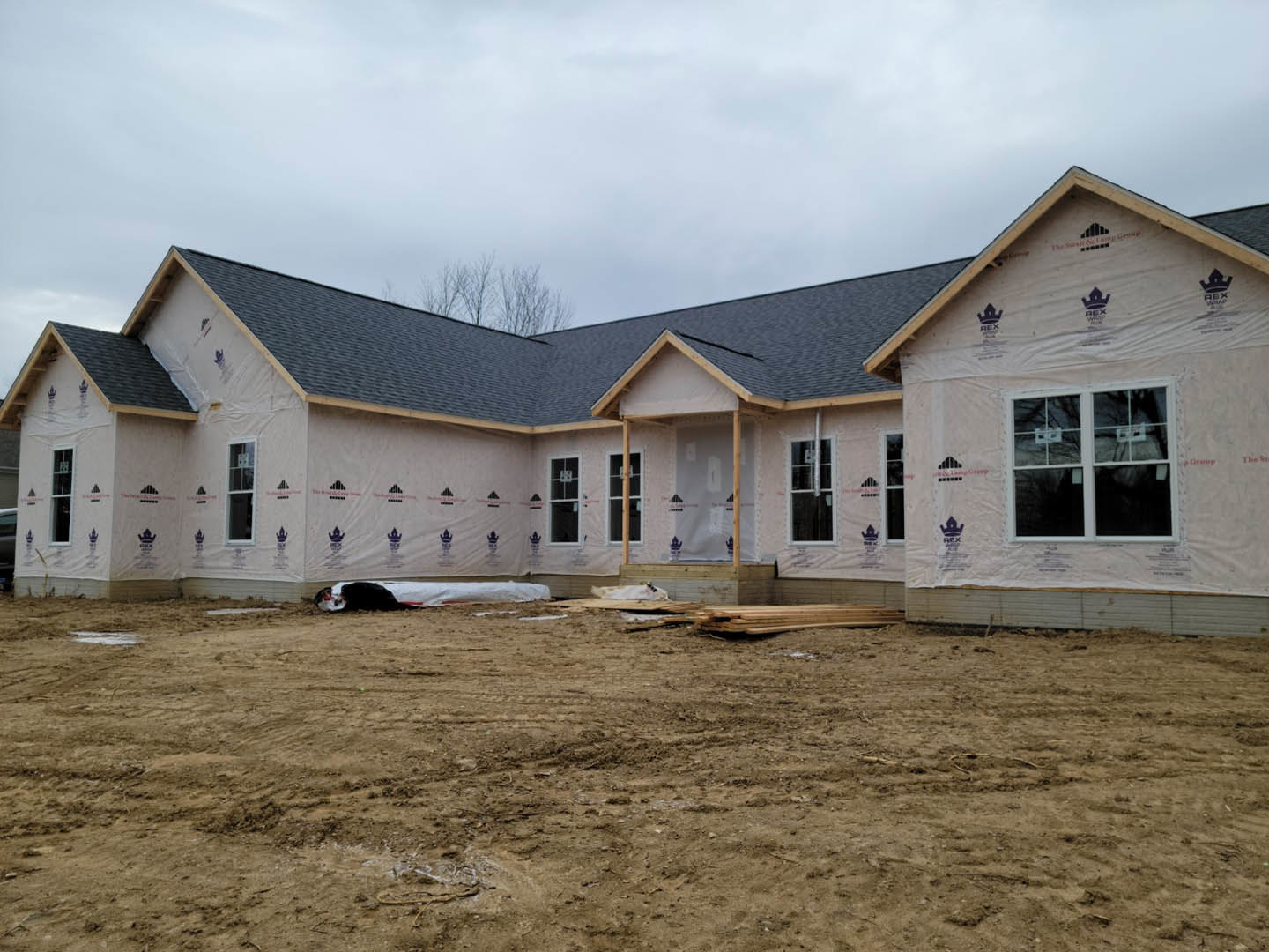 Two-story house under construction with exposed framing, unfinished porch, dirt field in foreground, piles of lumber, cloudy sky above