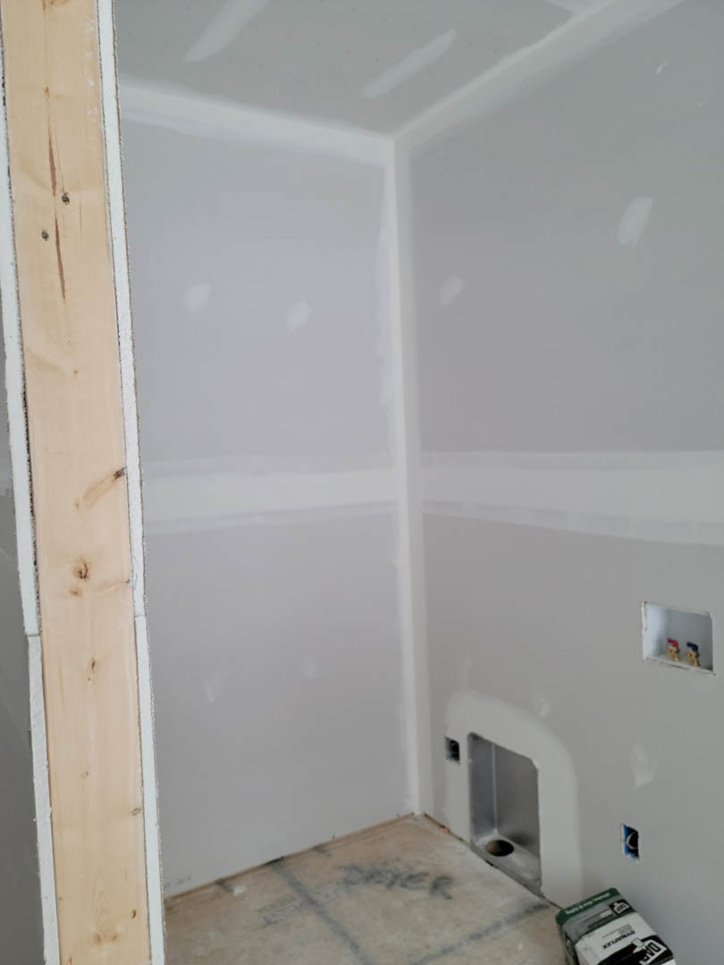 Drywall room under renovation with exposed hole in wall, white toilet, wood panel, white shelving with small toy figures, and construction materials visible