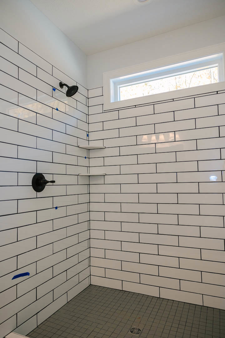Black shower head mounted on white tile wall, built-in shelves, and a window allowing natural light into the bathroom shower area