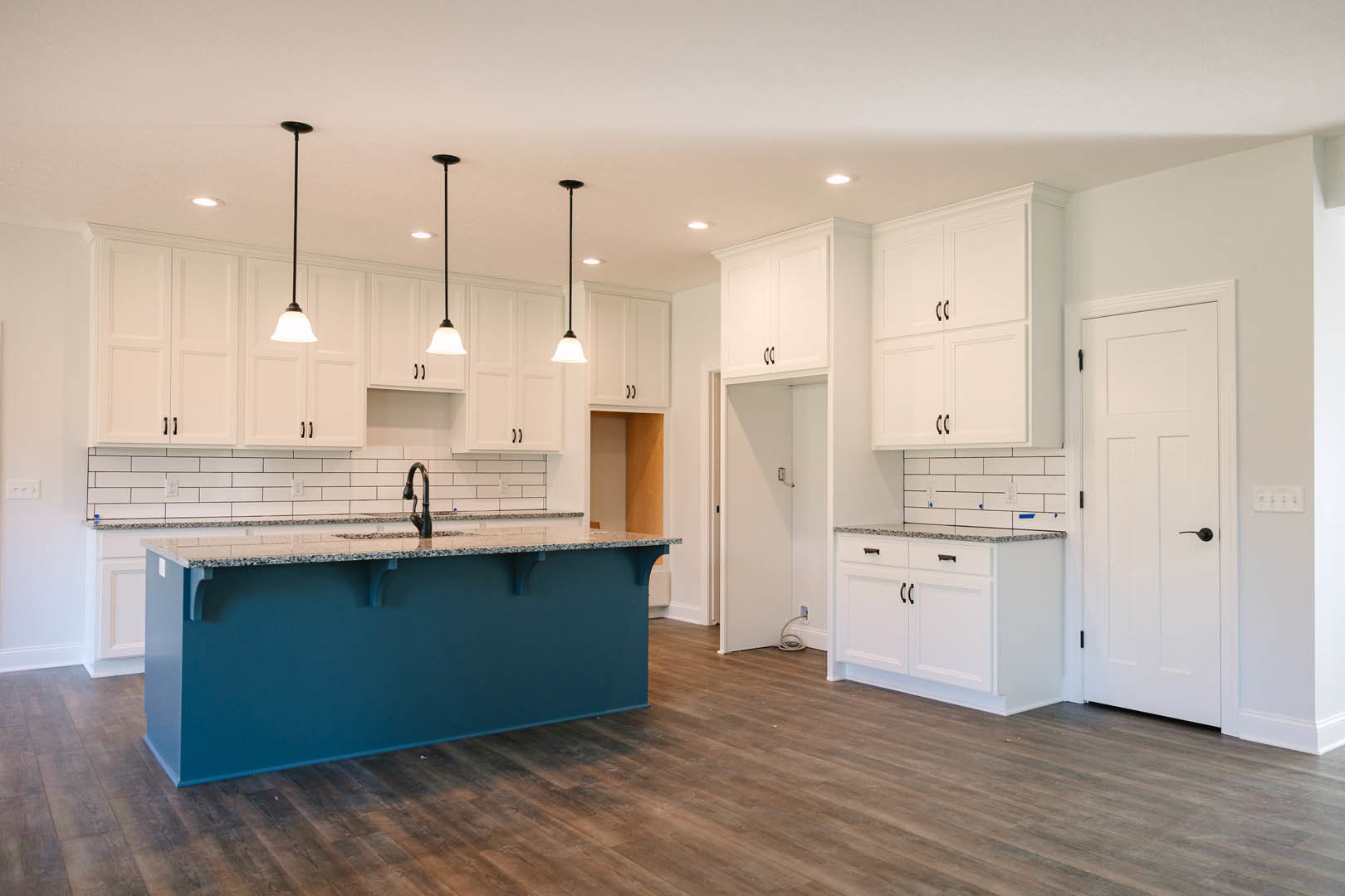 Blue kitchen island with marble countertop, white shaker cabinets with black handles, wood flooring, white door with black handle, and tiled backsplash