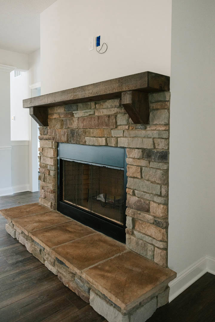 Stone fireplace with black metal frame and wire mesh screen, white mantel shelf above, blue painter’s tape on adjacent white wall, hardwood floor visible.