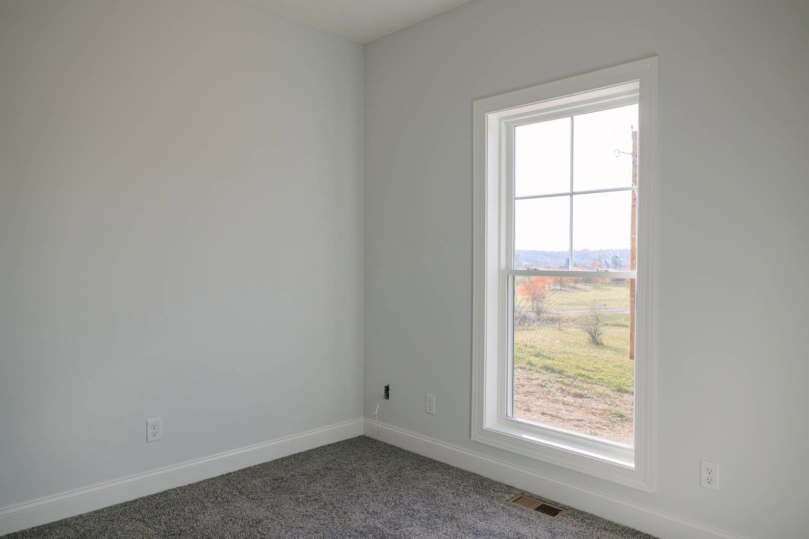 Carpeted floor with white baseboards, large window overlooking field and trees, metal floor vent, neutral plaster walls, window blind partially visible