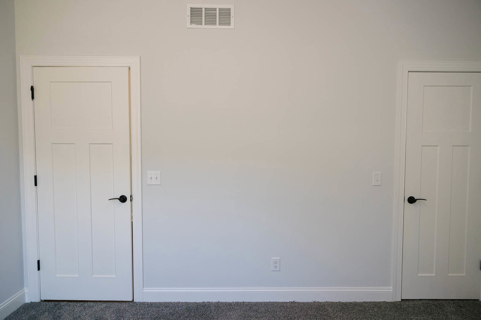 White paneled door with black handle, white wall vent, light switch, and beige carpet flooring in a residential interior.