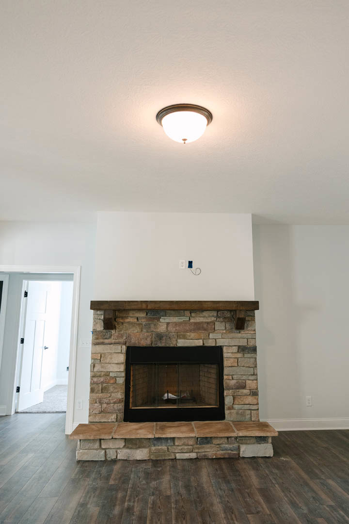 Brick fireplace with wood mantel, glass door, and hearth in a den with plaster walls, wood flooring, ceiling light fixture, and white door illuminated by natural light