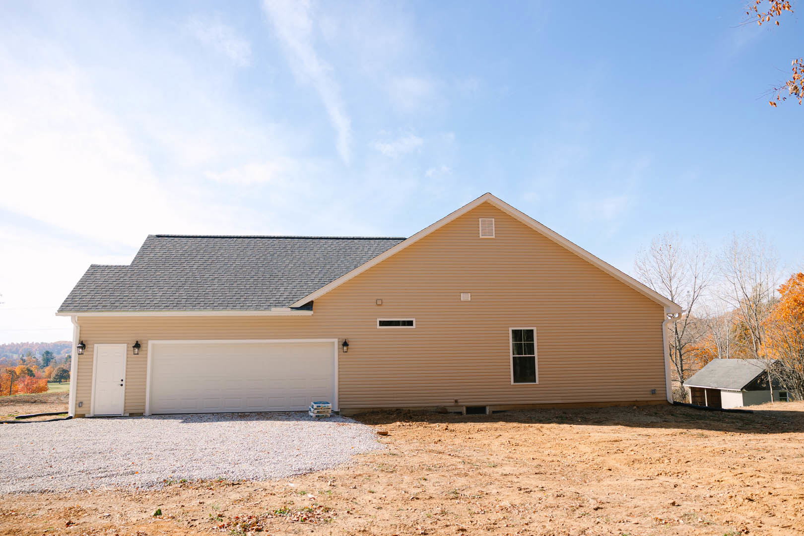 White cottage-style home with open front door, attached garage featuring paneled door, white-framed window, gravel driveway, and dirt ground surrounded by trees under partly cloudy