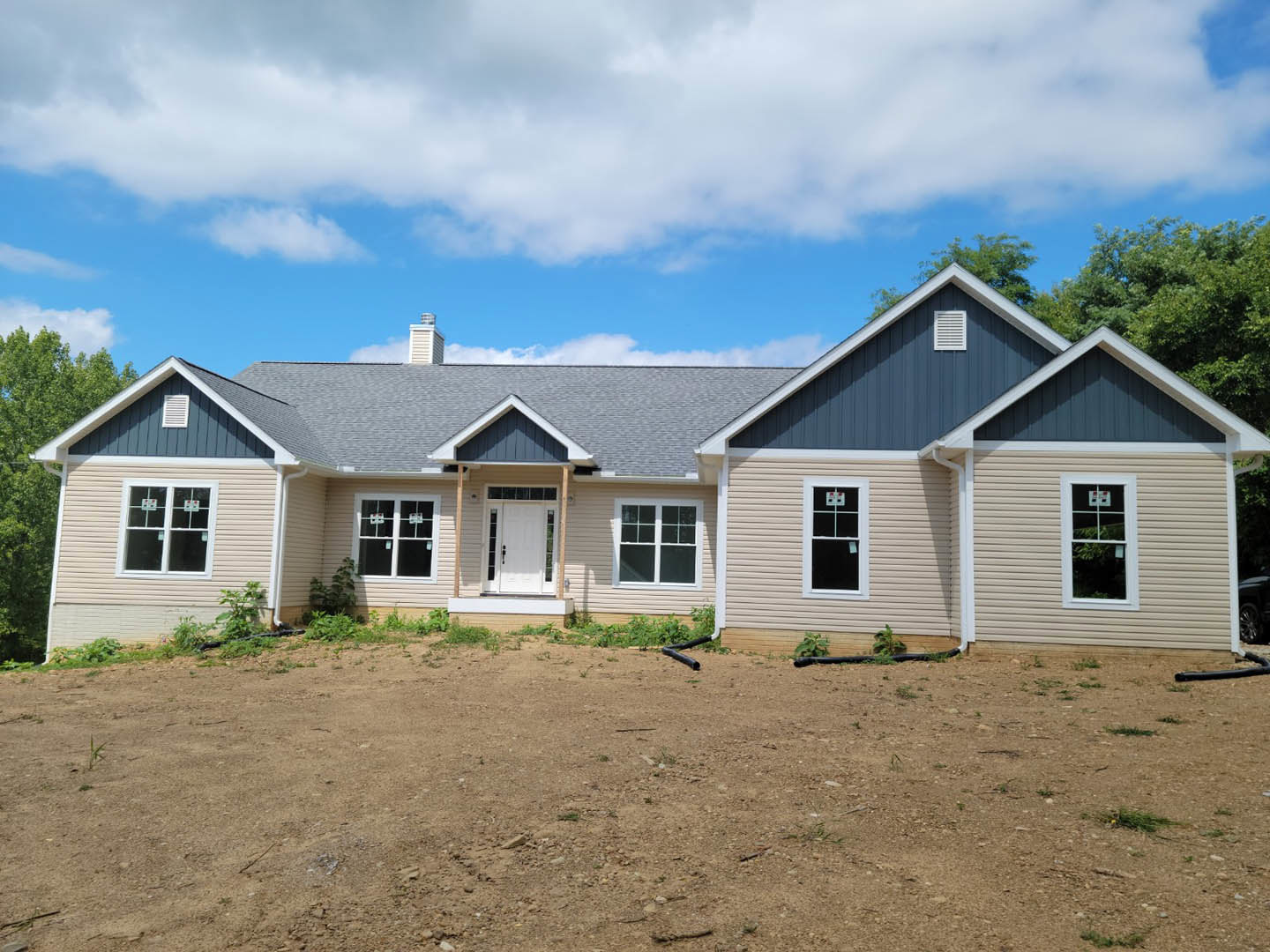 Two-story house under construction with white siding, white framed windows, and a white front door; basketball hoop visible in window; dirt yard with exposed pipe; trees in