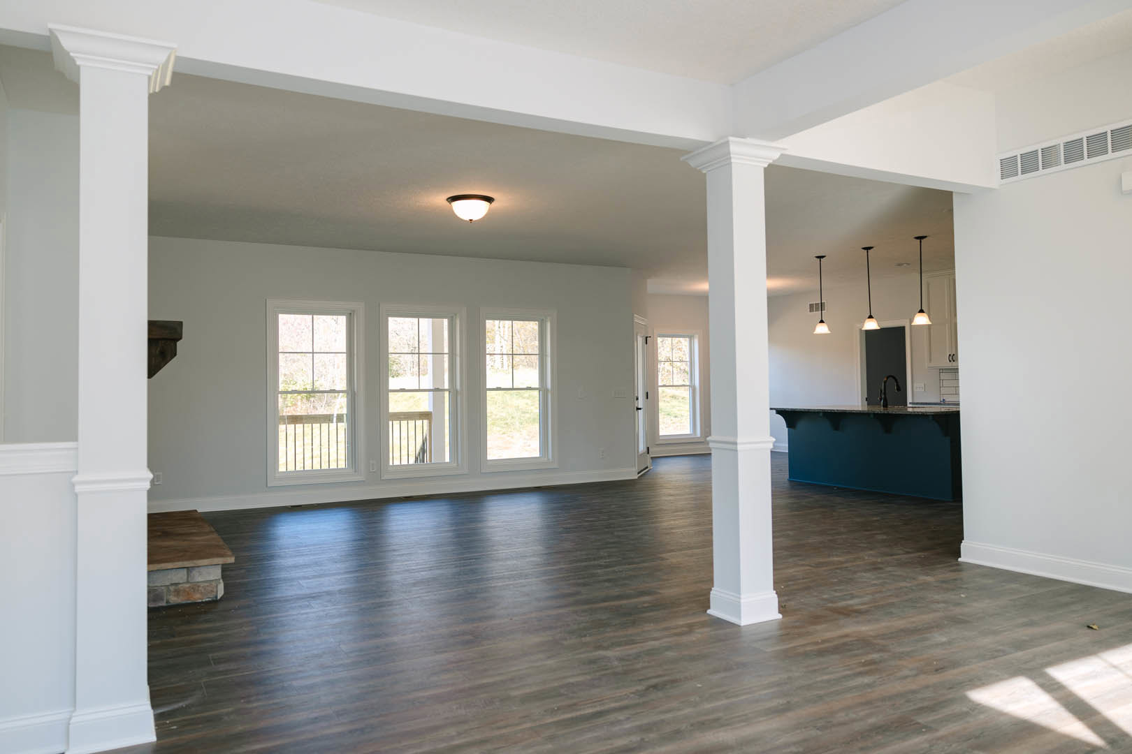 Spacious open floor plan featuring dark wood flooring, white columns, modern kitchen with light fixtures on the ceiling, large windows with exterior railings, and a close-up of a