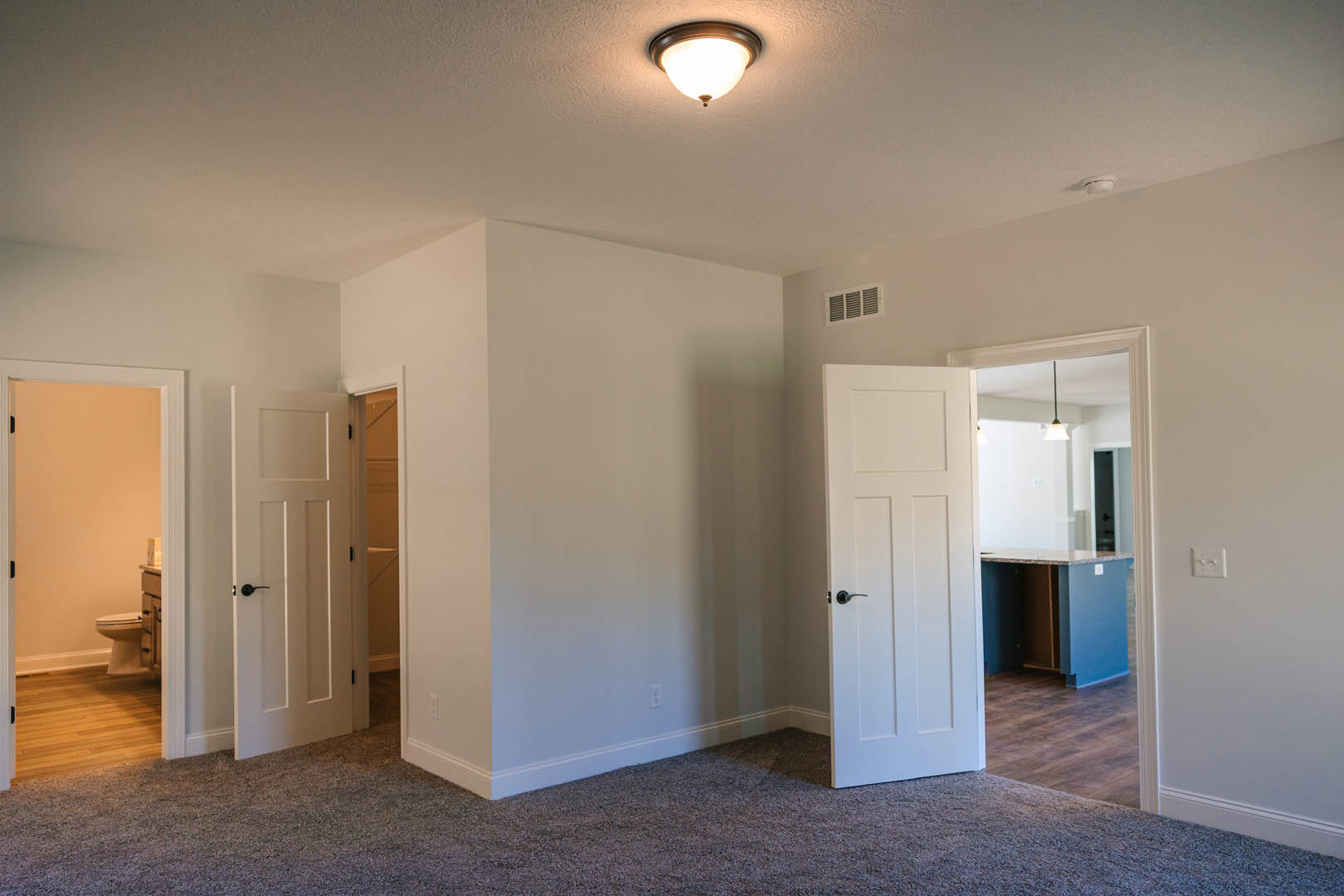 White-walled room with white doors featuring black handles, ceiling light fixture, visible bathroom with toilet and sink, wall vent, light-colored flooring