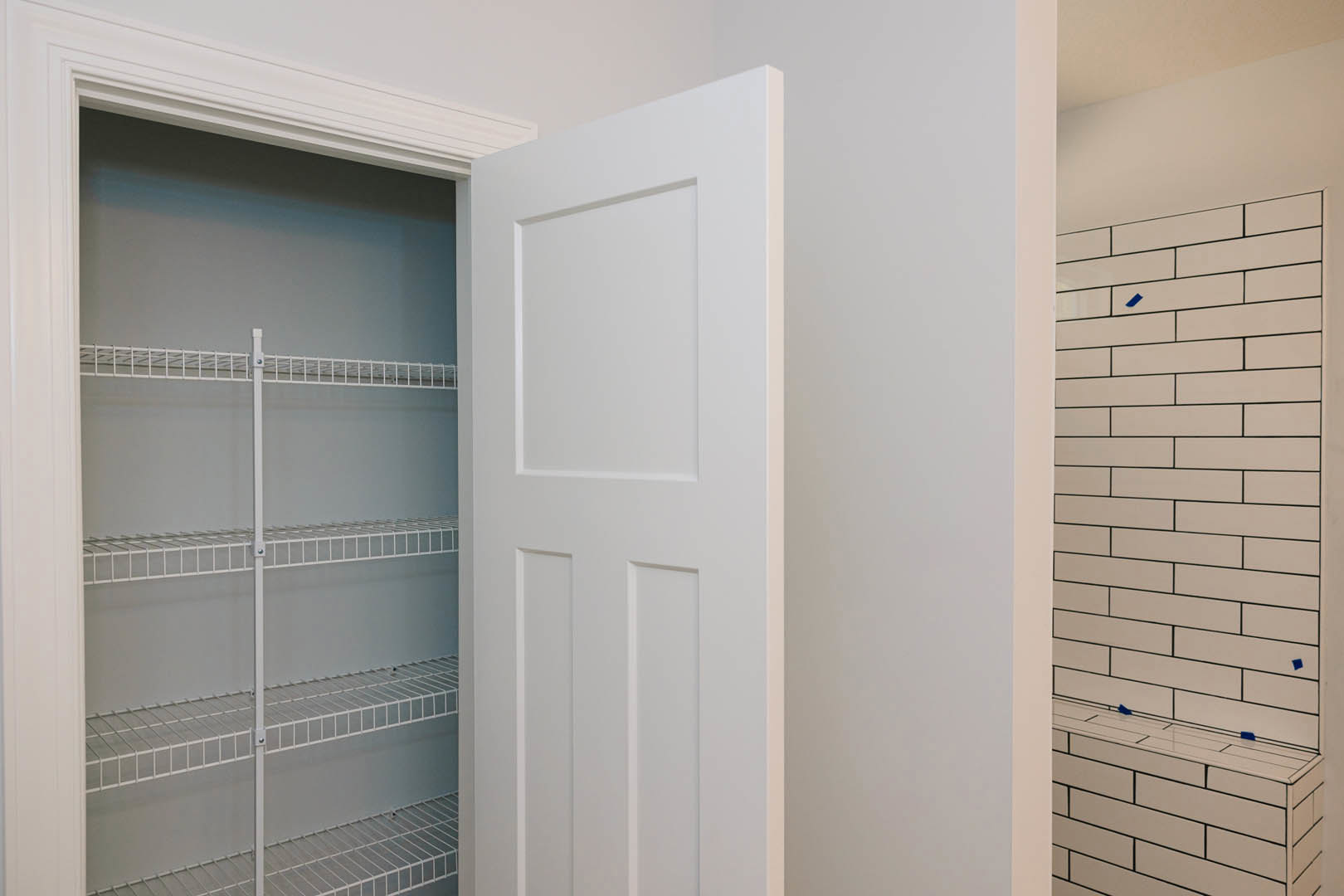 White paneled door open to a closet with white metal shelving, white walls, and rectangular tile flooring with blue square accents.