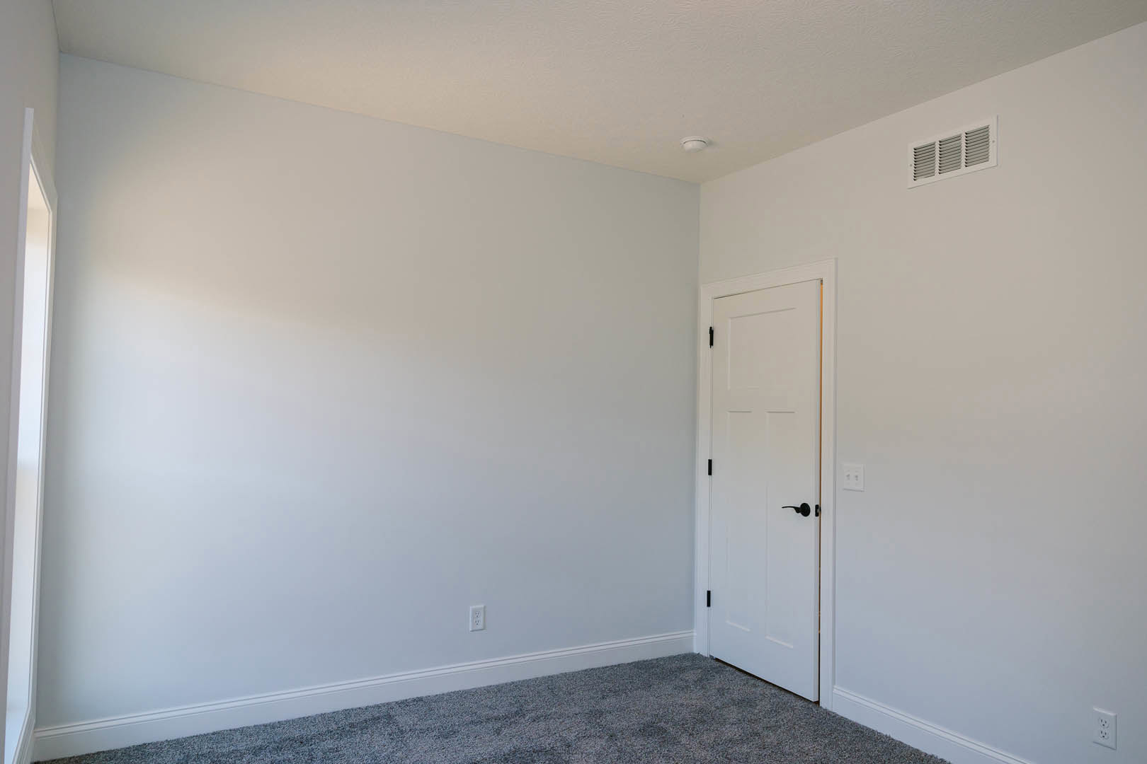 White paneled door with black handle, beige carpeted floor, white walls and ceiling, wall vent visible near baseboard