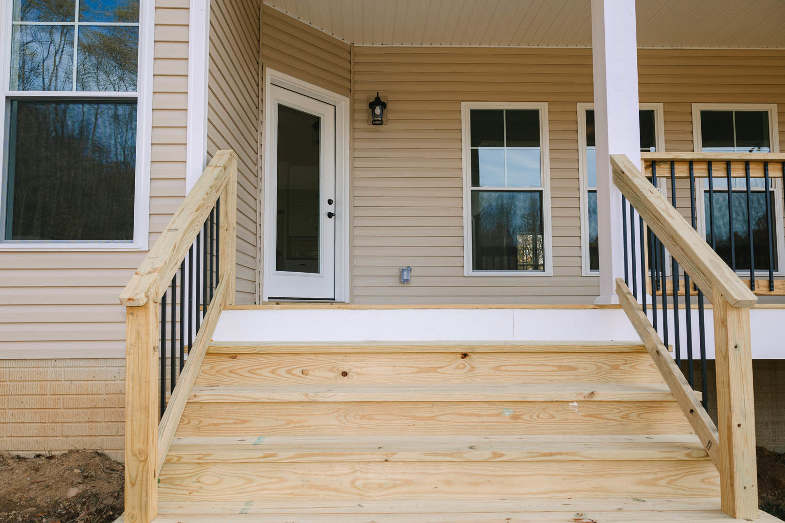 Wooden staircase with baluster and handrail leading to white door with glass panel, white-framed windows reflecting trees, brown-framed white ceiling, wood siding exterior