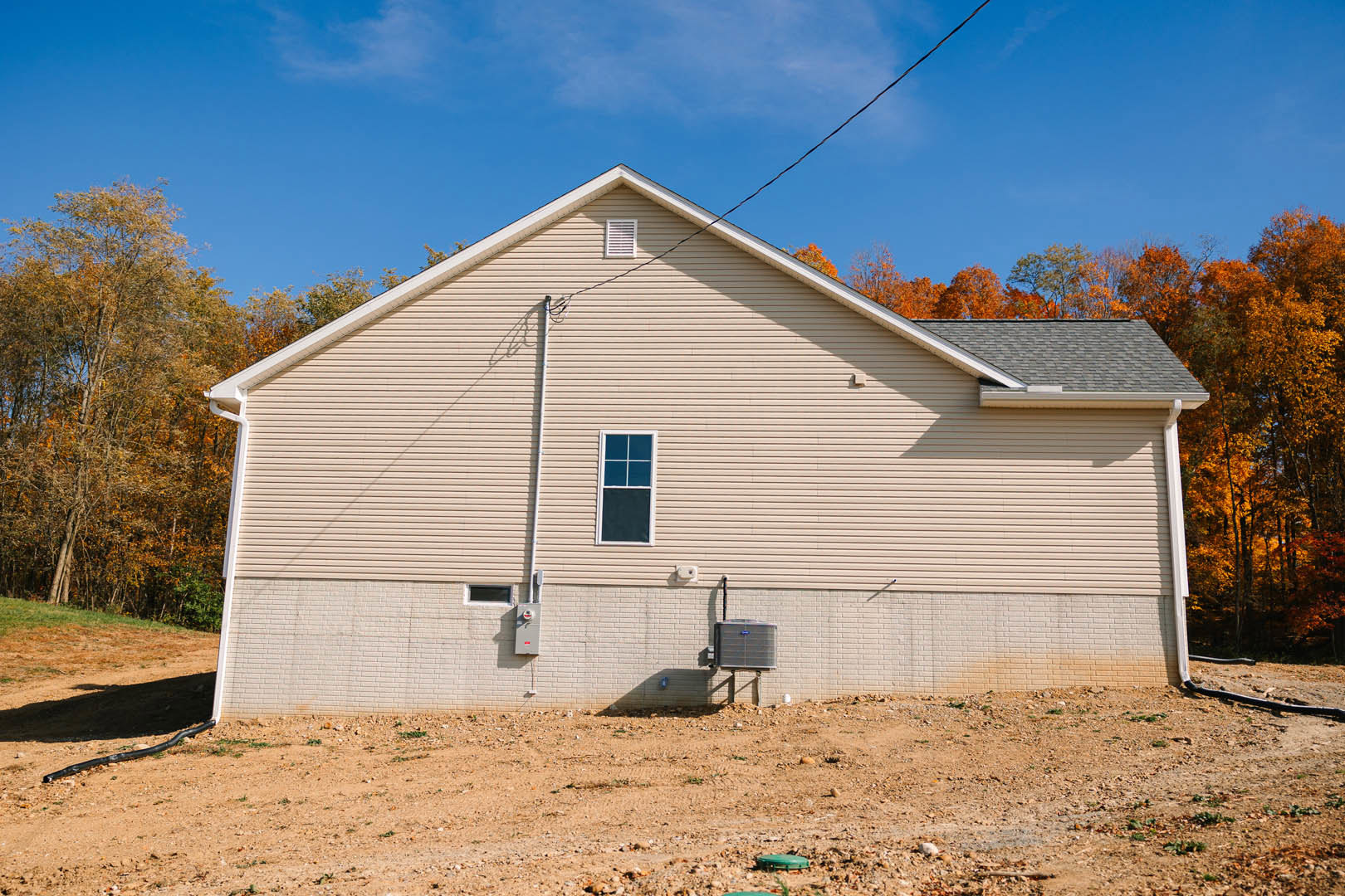 Two-story house with light-colored siding, black screened window, heat pump unit near foundation, power lines overhead, tree with yellow leaves, dirt yard, green object in