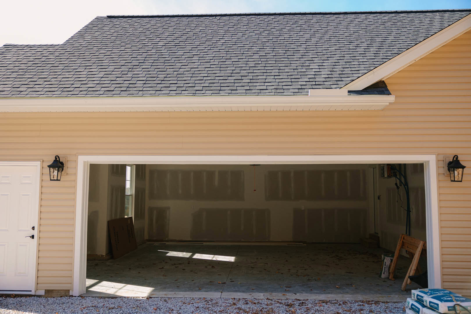 Detached garage with pitched roof, white paneled garage door featuring black handle, light-colored siding, ladder leaning against wooden structure, bag and stacked packages on