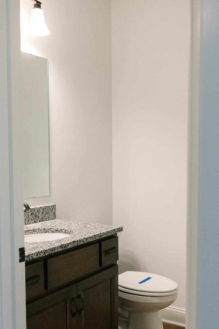 White bathroom with marble countertop, undermount sink, modern faucet, toilet featuring blue painter’s tape on lid, light gray tile flooring, and white shaker-style cabinet.