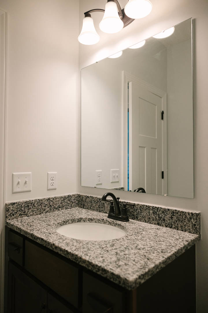 White ceramic vessel sink with chrome faucet set on quartz countertop, large frameless mirror above, gray tile backsplash, modern light fixture, and white cabinetry visible in
