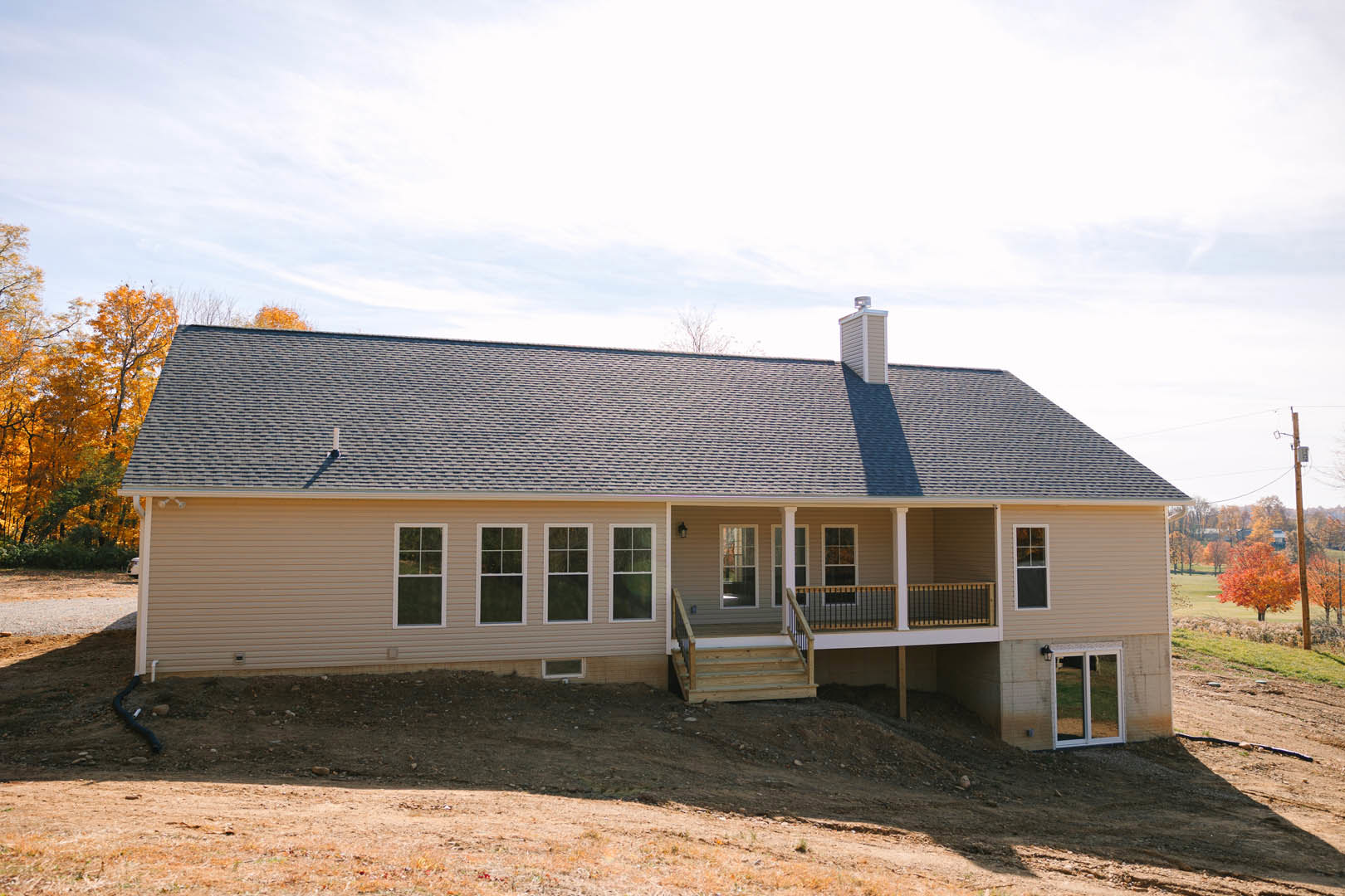 Two-story home with wide covered front porch, wooden stairs, white-framed windows, gray roof, and tree with orange leaves in yard