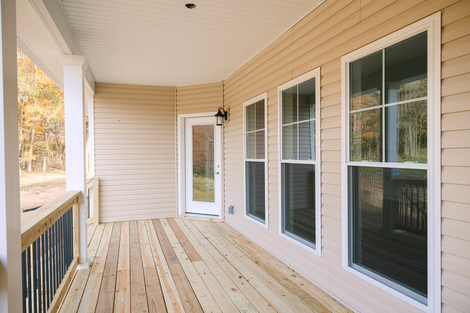 Wooden porch with glass door, multiple windows, white ceiling, and wood railing, outdoor lamp mounted near entry.
