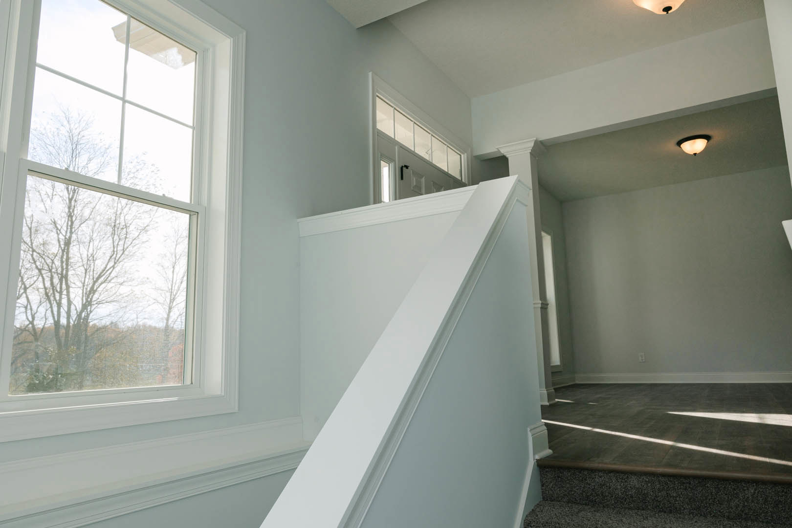 White staircase with matching walls, tile floor illuminated by ceiling light fixture, large window framing leafy trees, white door topped with transom windows.