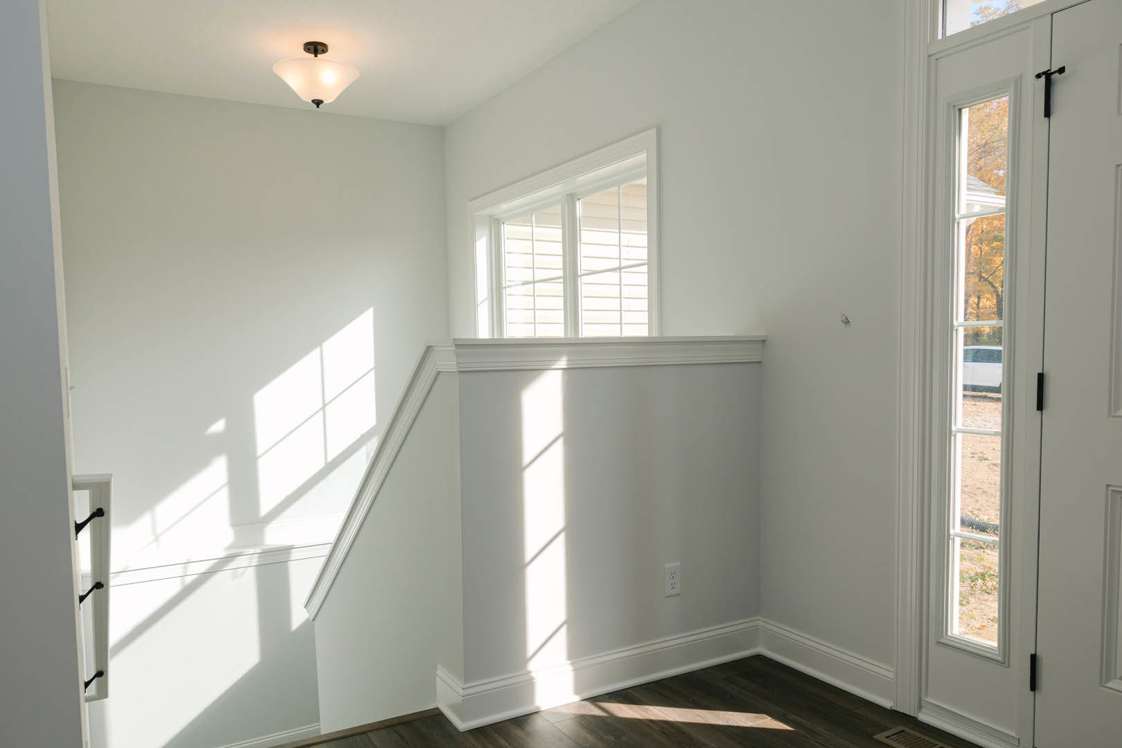 Bright white room featuring a window with blinds, staircase with white railing, ceiling light fixture, and a glimpse of a white car outside.