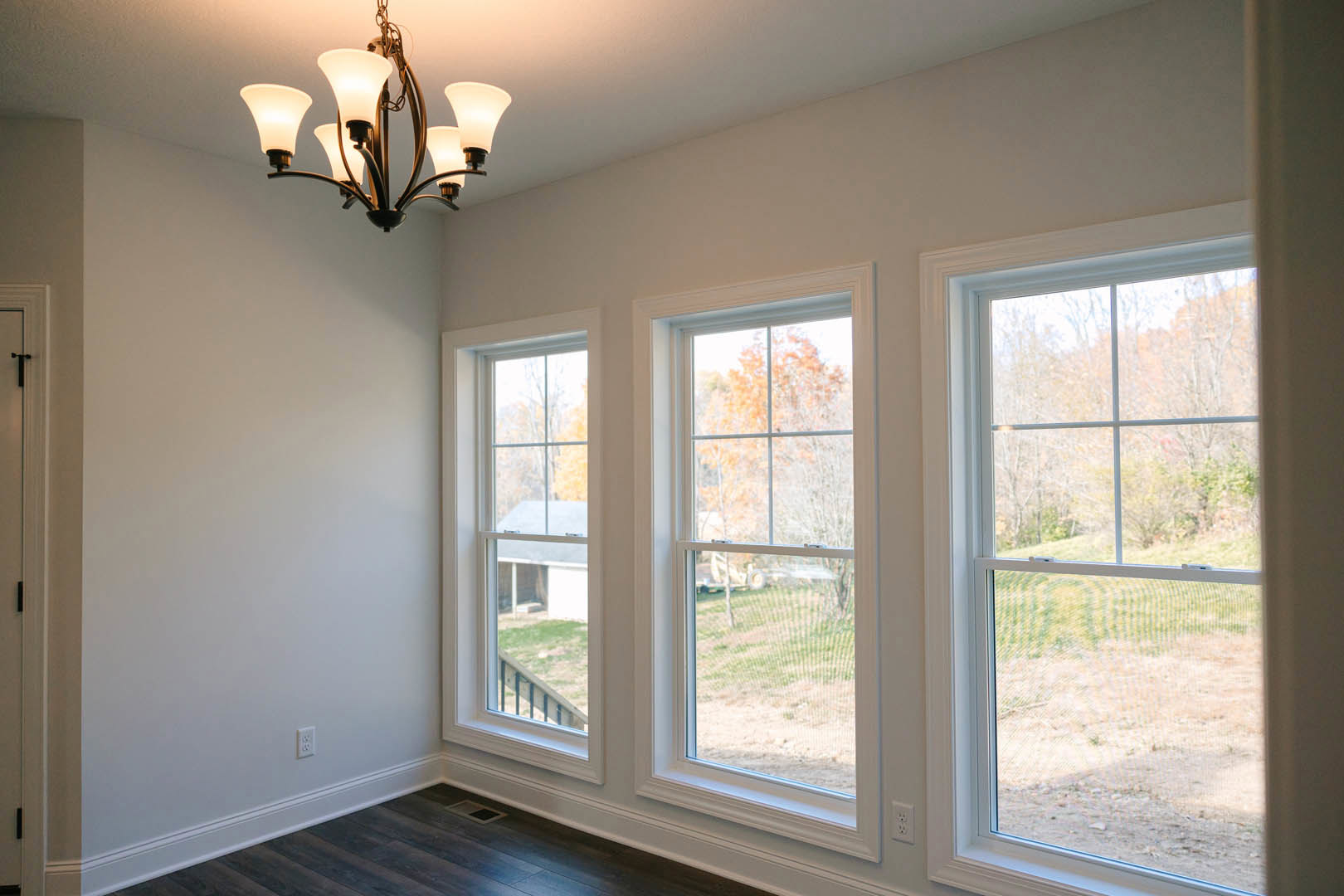 Bright room with three large windows, white walls, crown molding, five-light chandelier, window views of green lawn and trees, ceiling vent, and window blinds