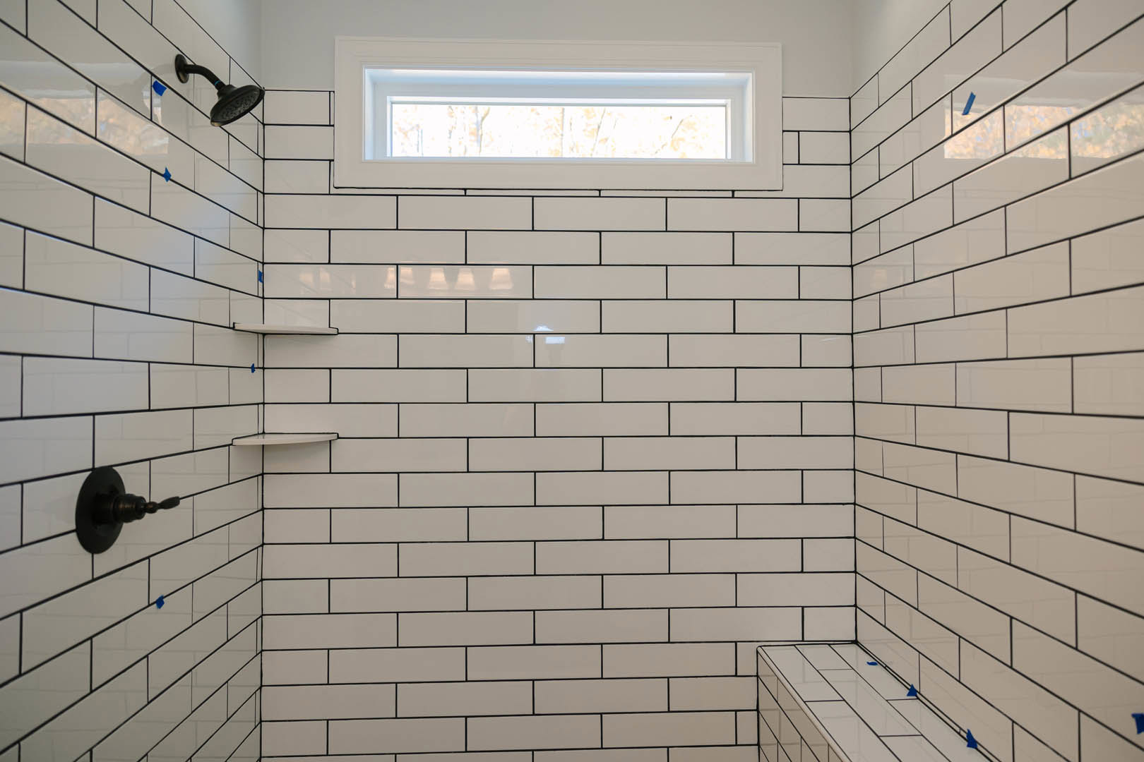 White-tiled shower with chrome shower head and rectangular window, black light fixture mounted on adjacent wall