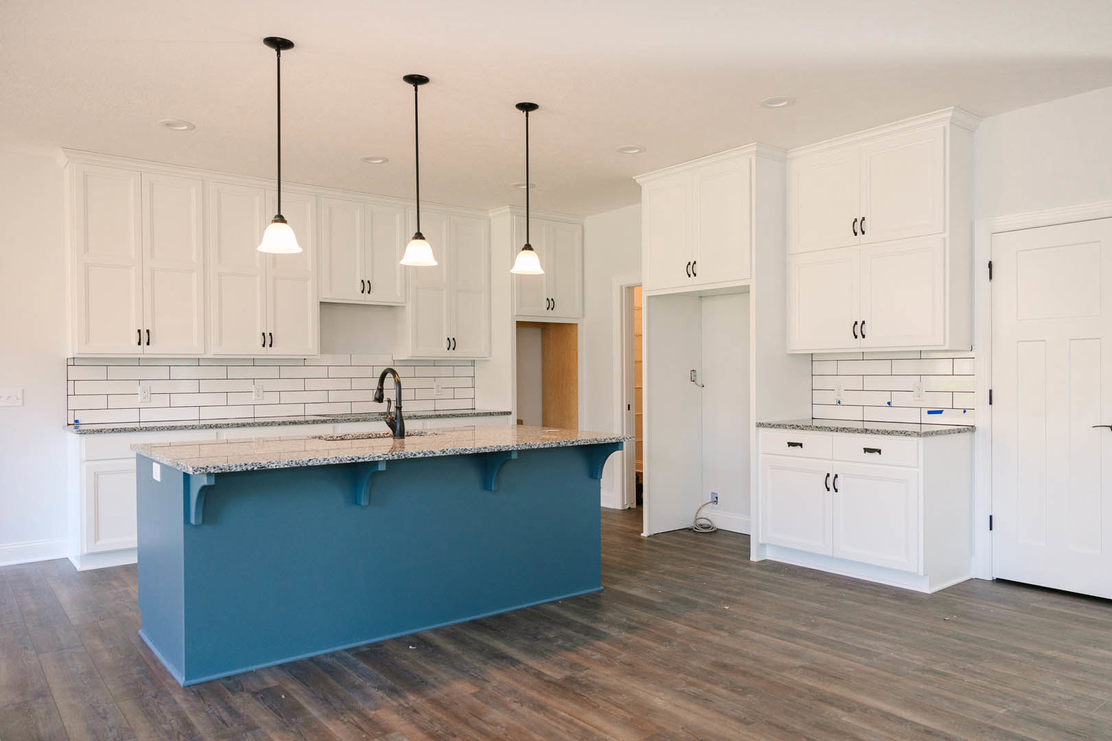 Blue kitchen island with marble countertop, white cabinets and drawers with black handles, white door with black handle, light-colored flooring, white hose on floor, white walls