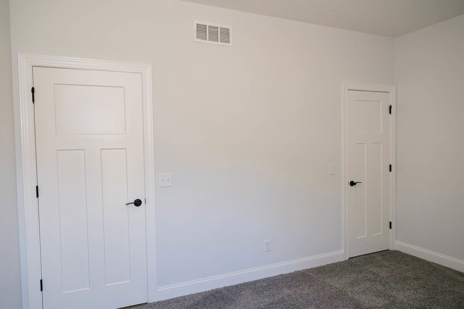 Carpeted room with two white doors featuring black handles, white walls, and ceiling vent