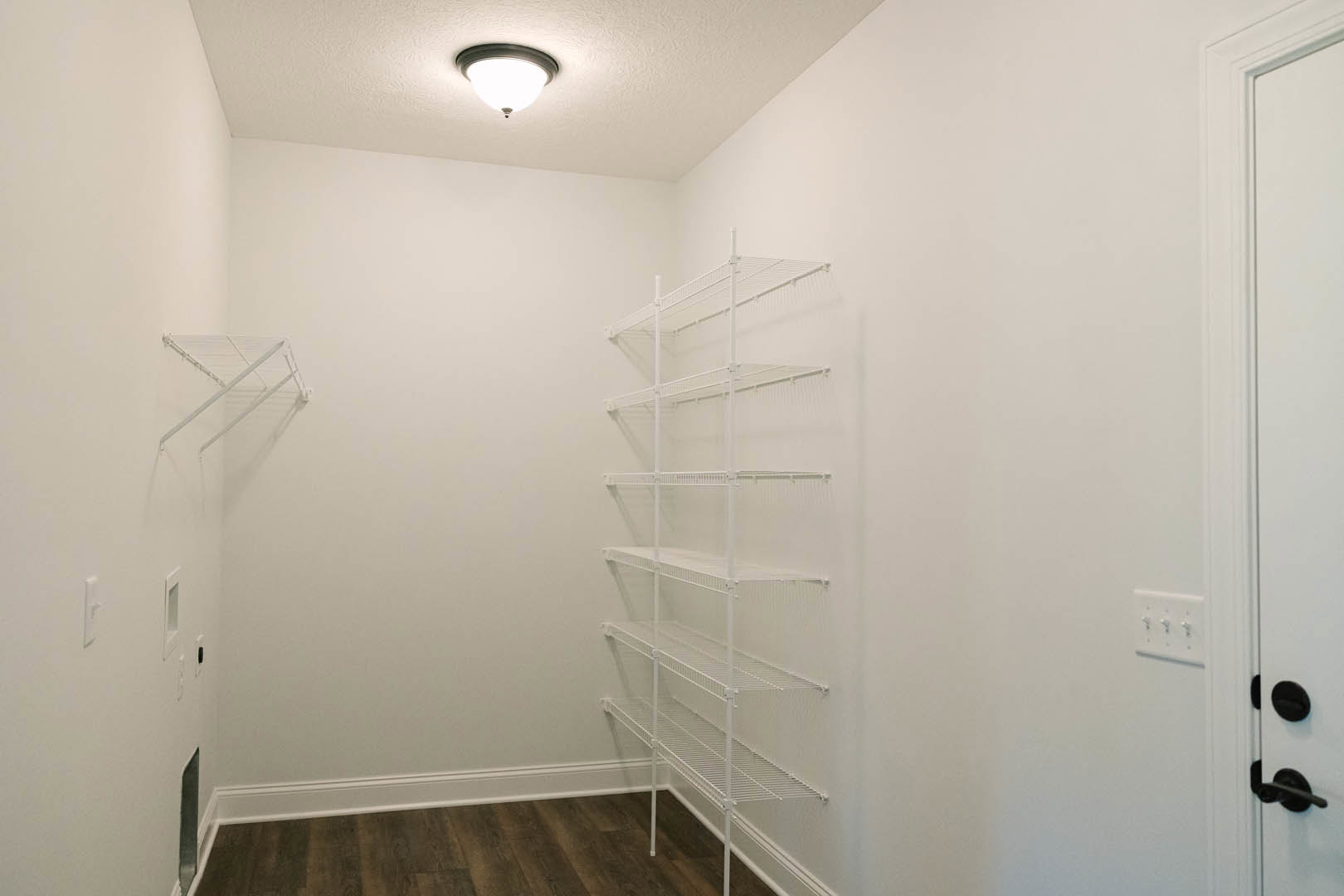 White wire shelving unit mounted on plaster wall beside white door, ceiling light fixture overhead, light switch visible on wall, hardwood floor below.