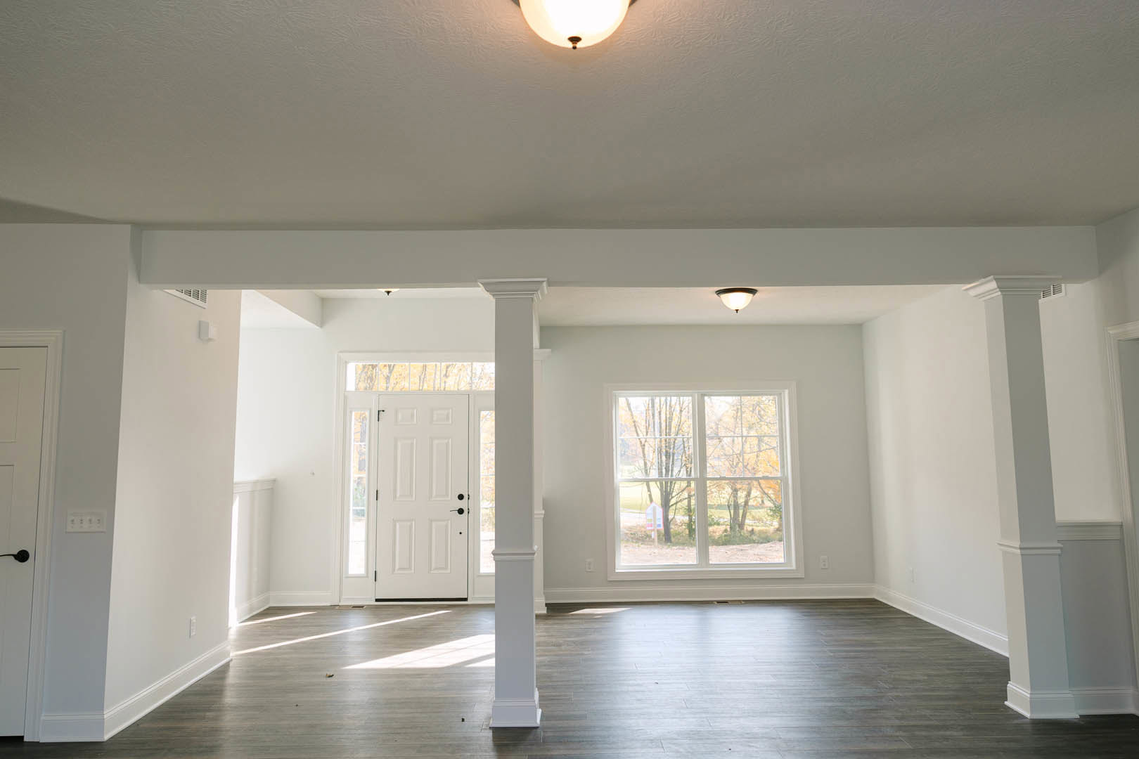 White column beside a white door with black knobs, wide window showing trees outside, light fixture overhead, wooden floor, white plaster walls with molding.