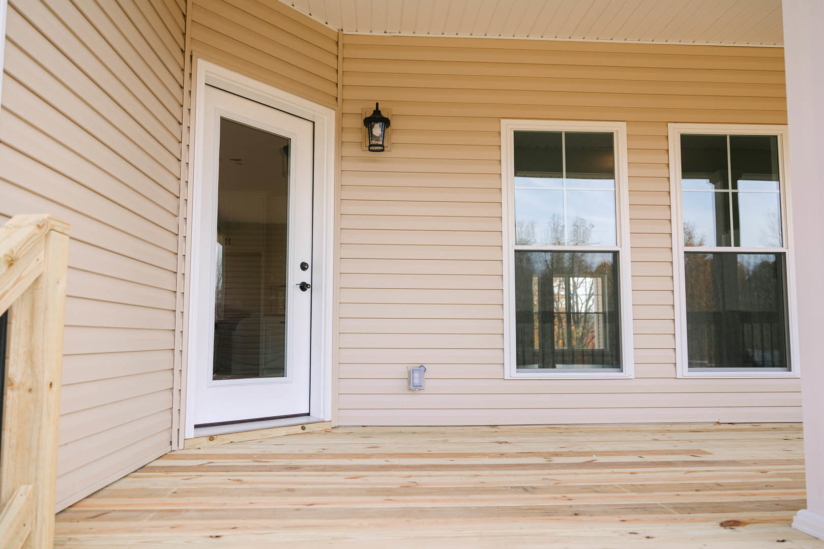 Front porch with wood plank flooring, glass door, large window reflecting trees, and wall-mounted light fixture on white siding exterior
