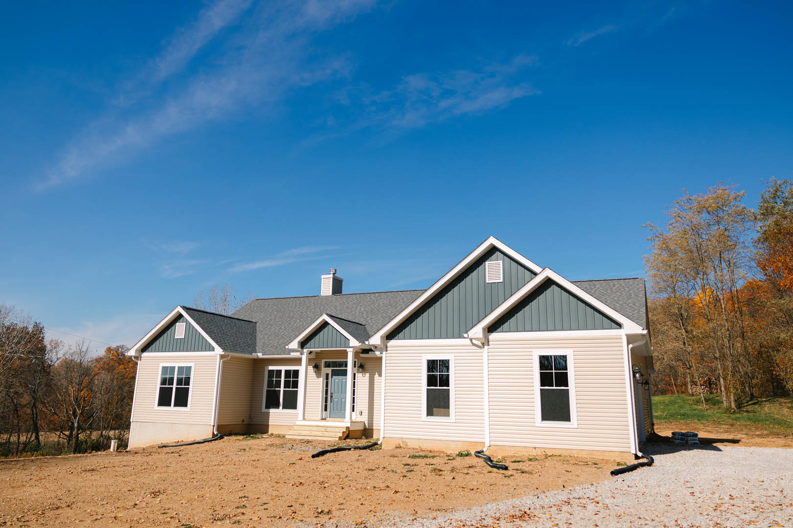 Gravel driveway leading to a cottage-style home with a blue door, white-framed windows, and orange-leaved trees under a clear blue sky