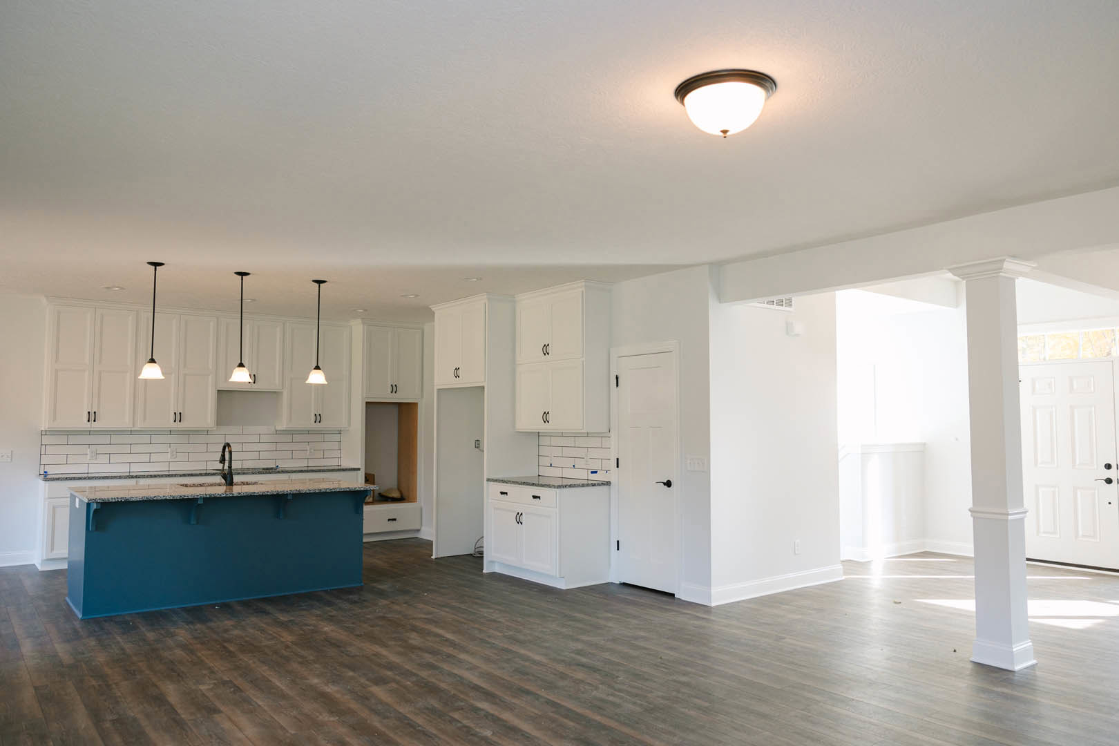 Open-concept kitchen and living room featuring wood flooring, white cabinetry with black handles, blue countertop, modern light fixture, and white door with black hardware