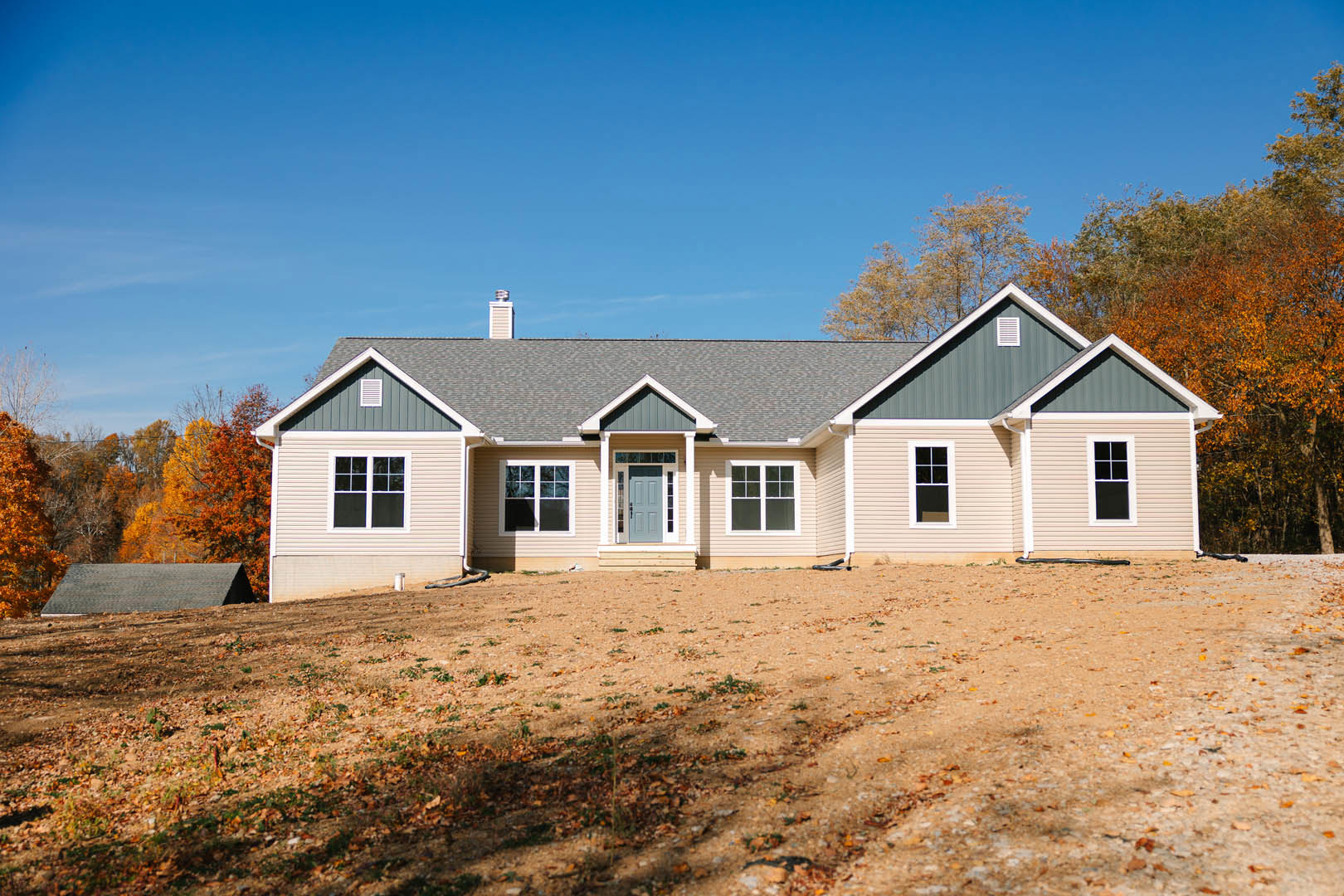 Two-story house with white-framed windows, blue front door, and gabled roof, set on a dirt patch with trees and a grassy hill in the background under a clear blue sky