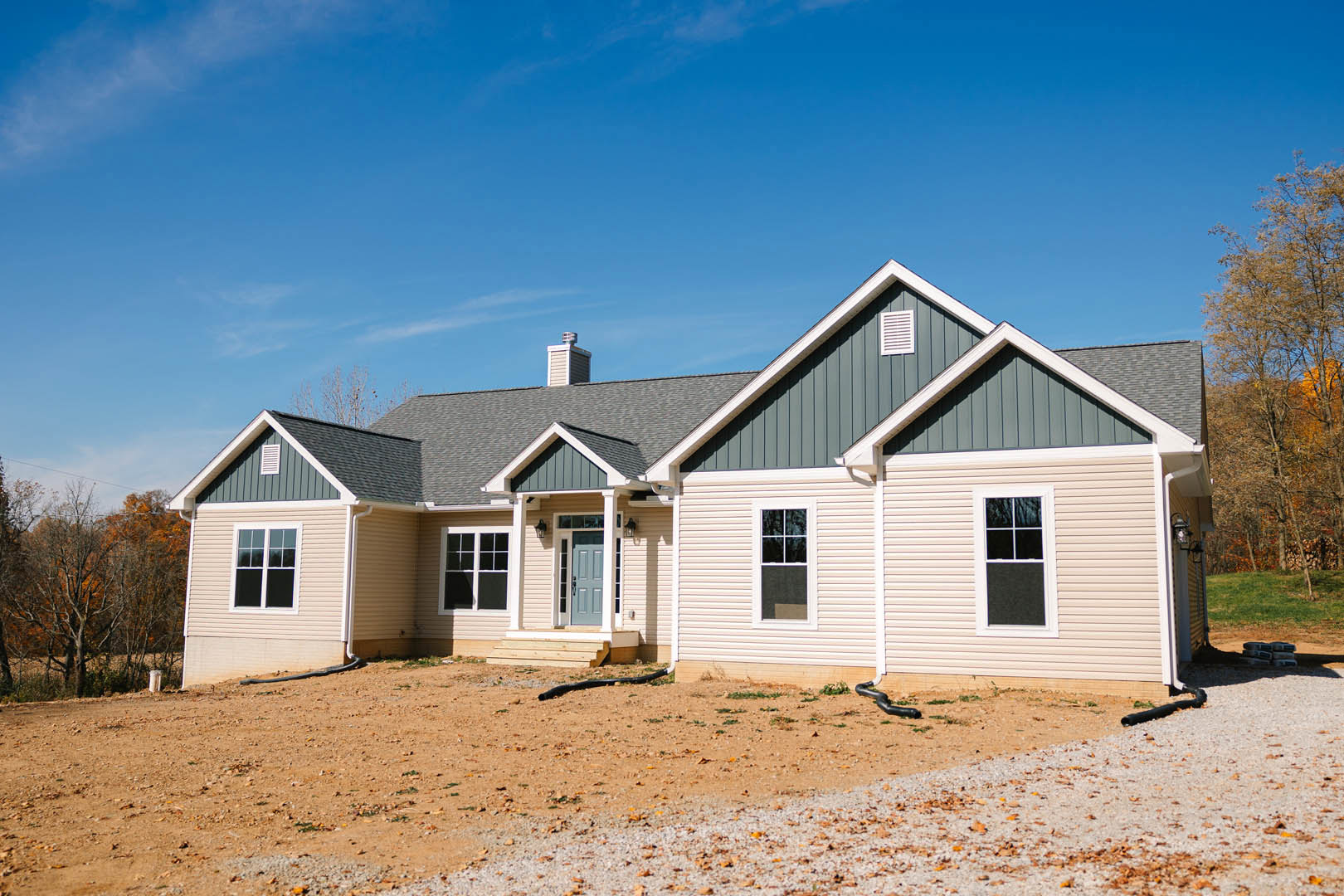 Single-story house with blue front door, white-framed windows, exterior wall vent, exposed pipe, and dirt yard under partly cloudy sky.