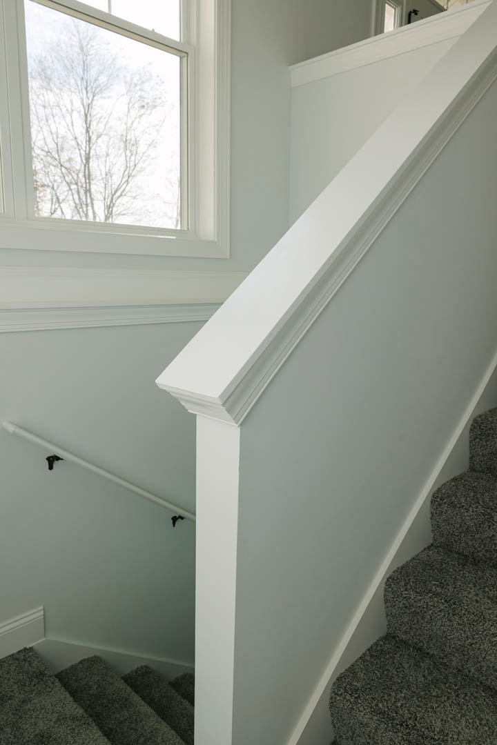 Carpeted staircase with wooden handrail, large window framing leafy trees, white plaster walls, black metal pipe, and natural daylight illuminating the space