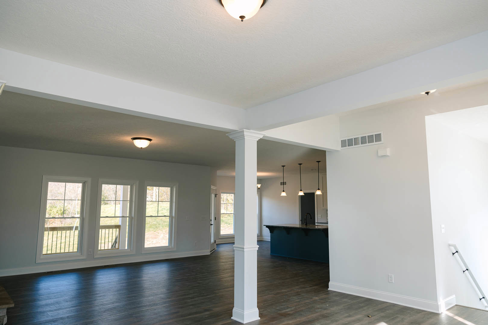 Open kitchen with wood flooring, white pillar, white walls featuring black accents, ceiling light fixture illuminated, large windows overlooking trees and grassy field.