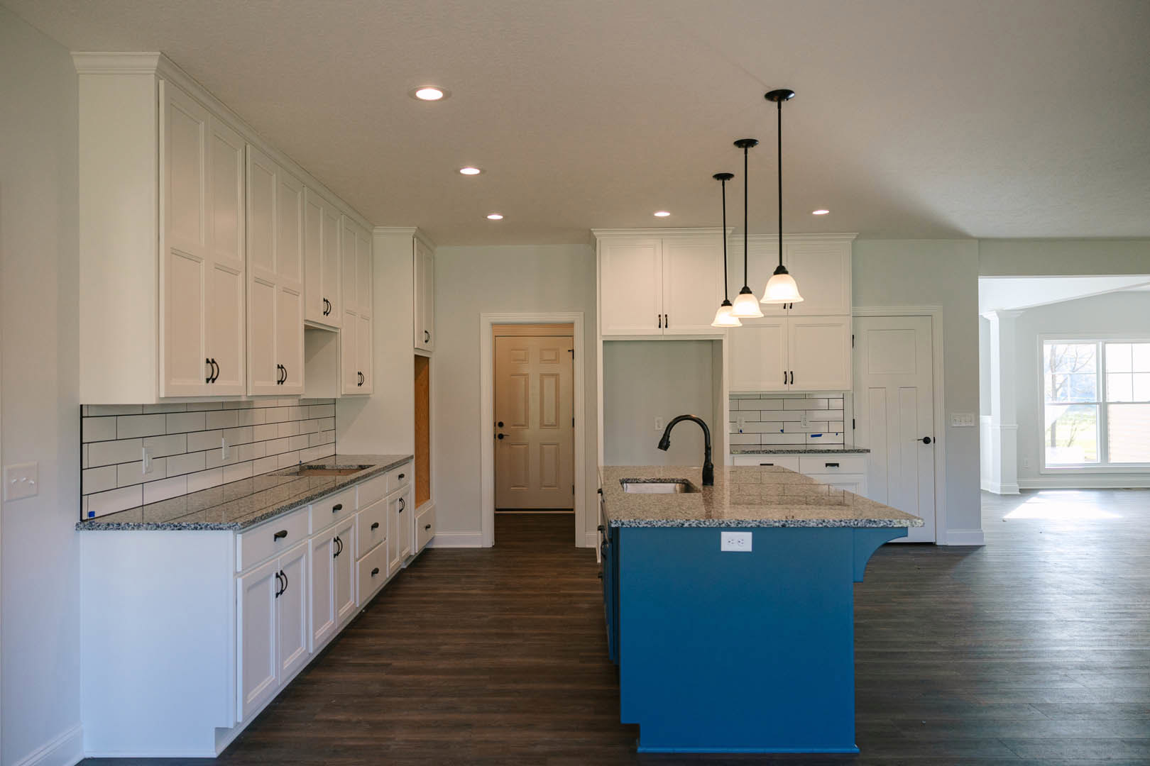 Blue kitchen island with granite countertop, surrounded by white cabinets and tile flooring, stainless steel sink, and a window letting in natural light.