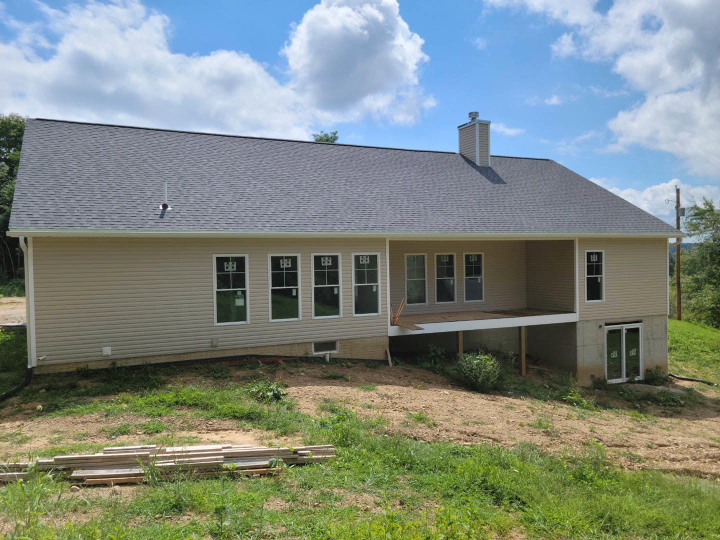 Two-story house with white-framed windows, brick chimney, covered porch, grassy yard, and a pile of firewood near the hill.