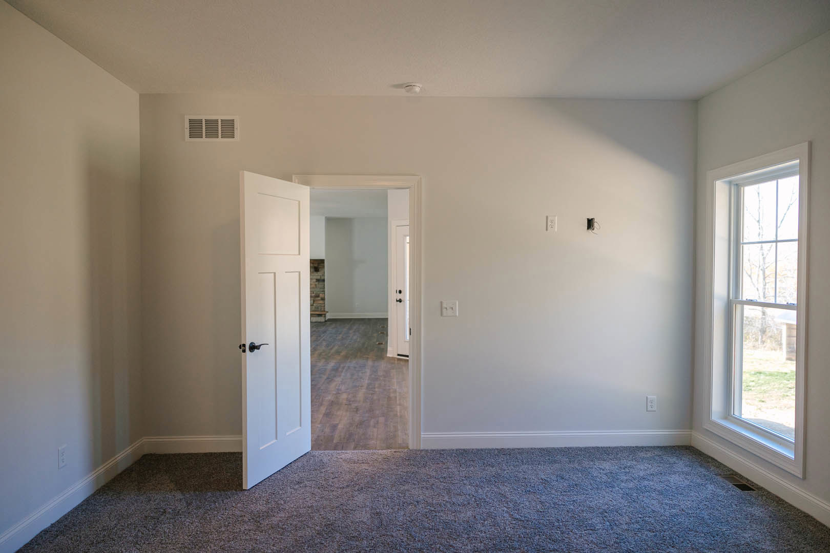 White-walled room with open paneled door, beige carpet flooring, large window framing leafy trees, decorative molding, and ceiling vent visible